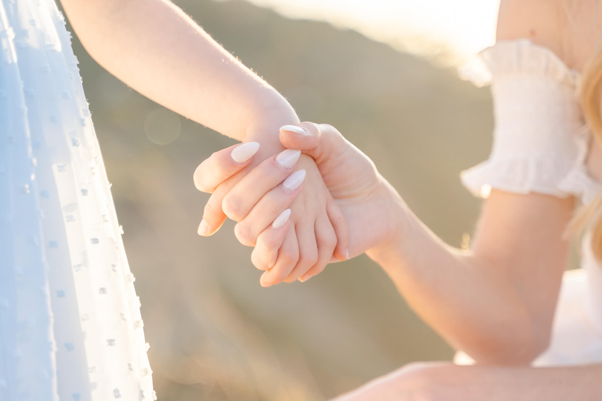 Mom wearing white with white nails holding hands with young daughter in blue dress. Close up photo of hands