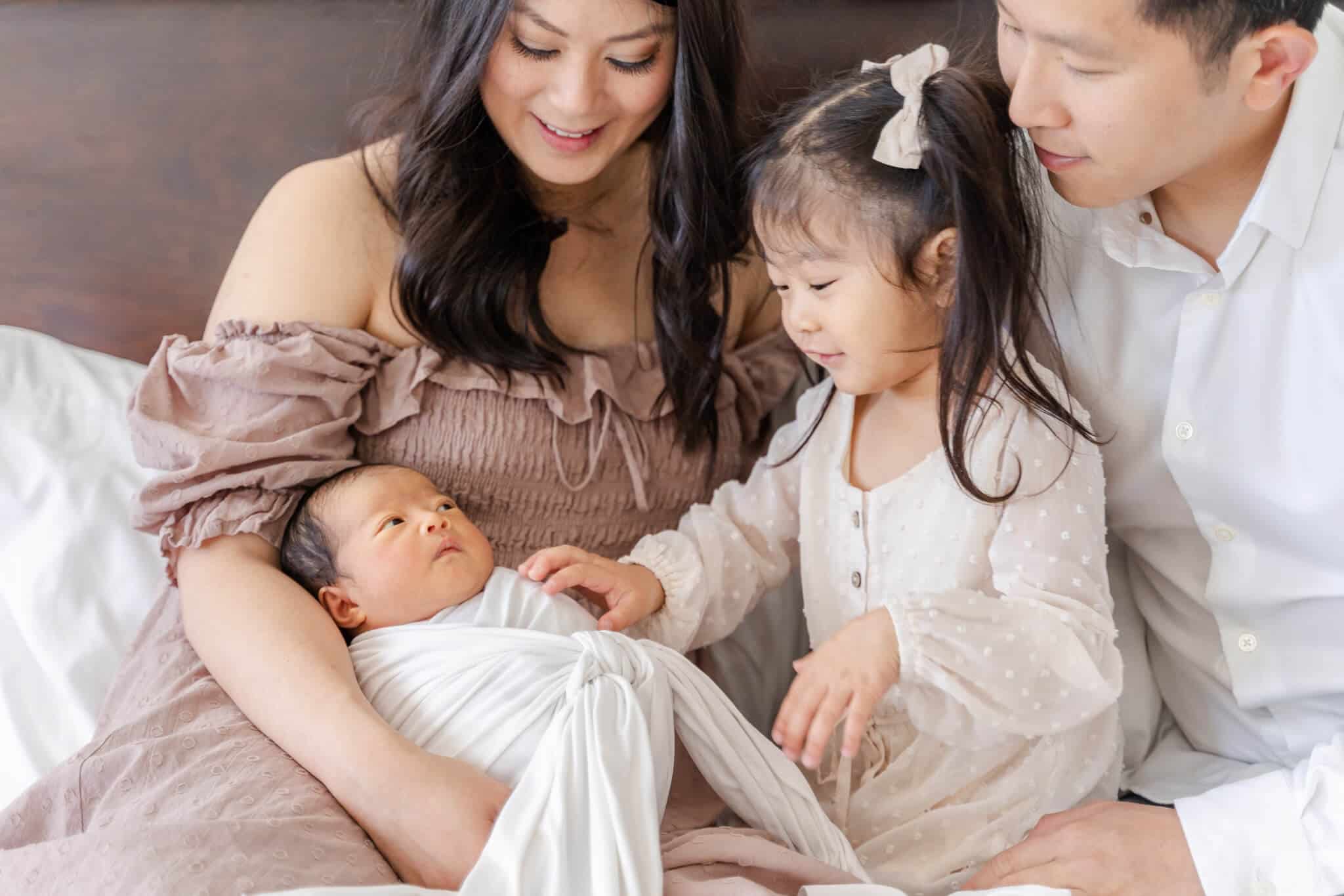 Asian family sitting on bed with toddler sister touching her baby sister- Irvine Pediatricians