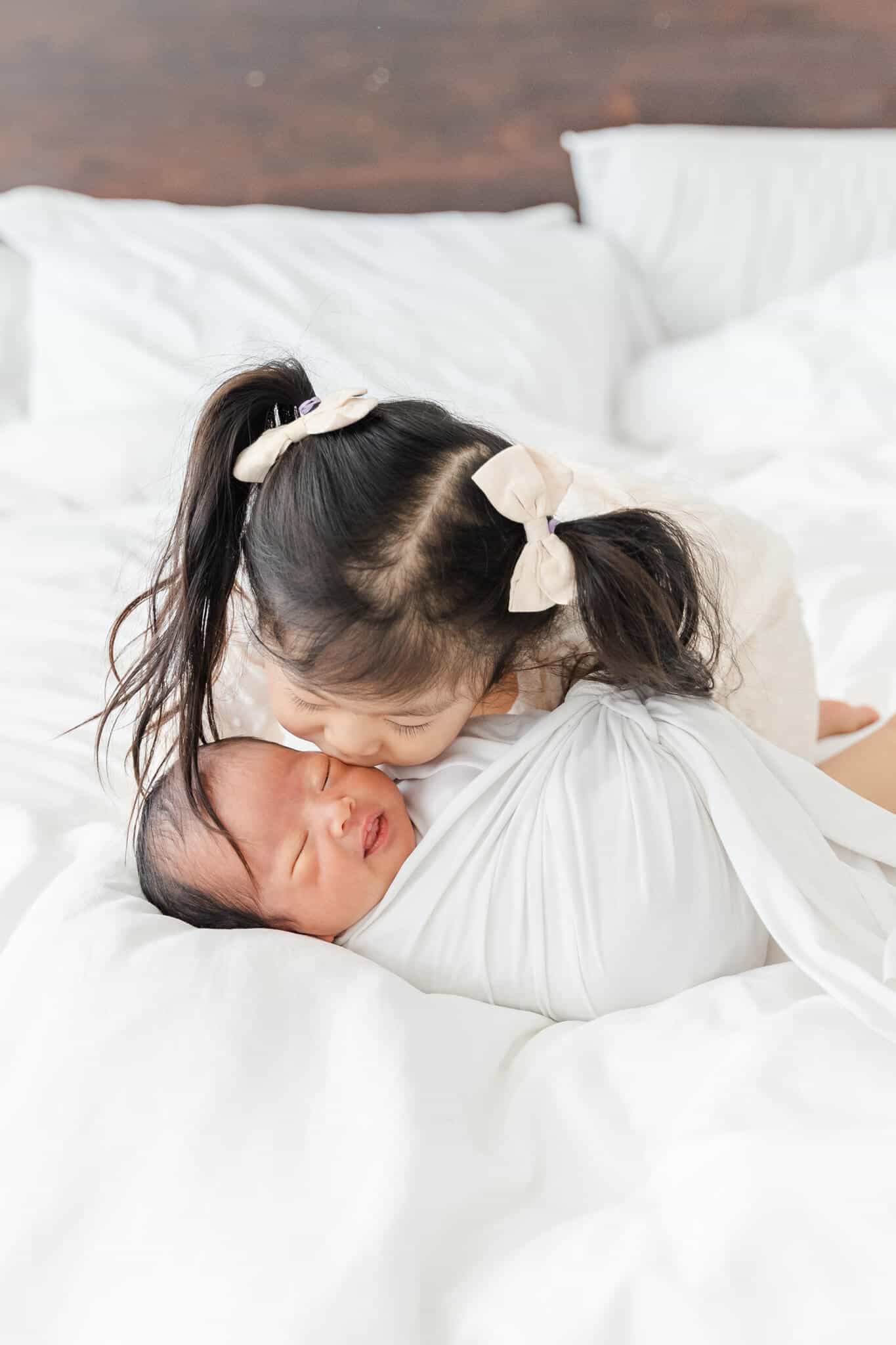 Asian toddler girl kissing her newborn baby sister on bed on a bed
