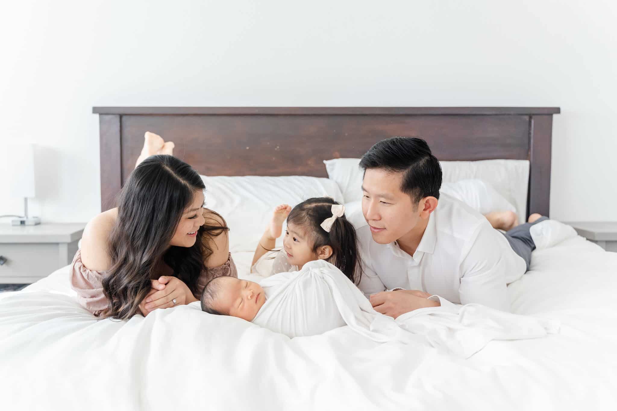 Family of four lying on bed and looking at newborn baby in light and airy room