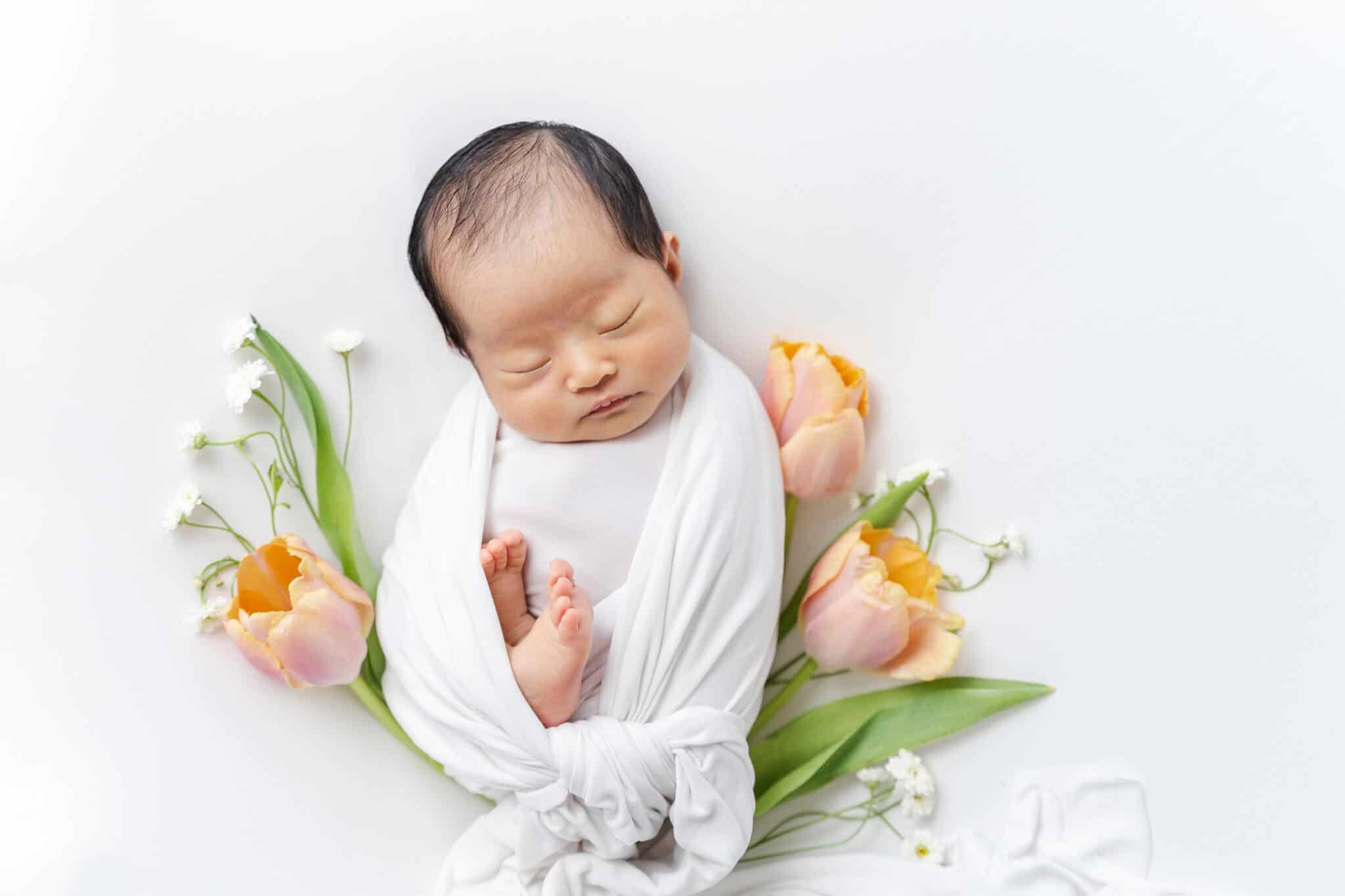 Sleeping newborn baby in white swaddle wrap surrounded by orange pink tulips and white daisies- Irvine Pediatricians