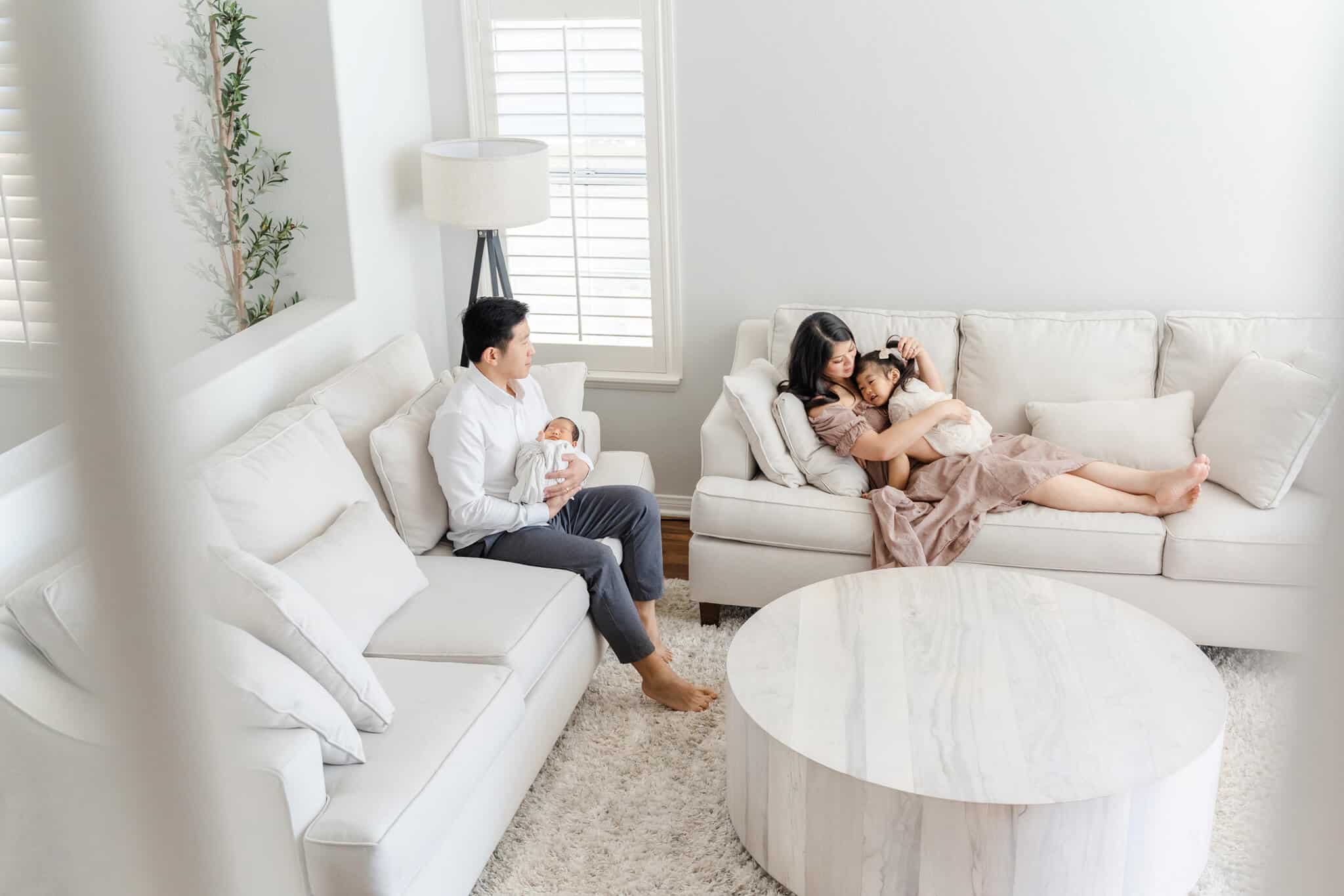 Mom cuddling with daughter, dad holding baby infant, relaxing on sofas in their light and airy home in the living room- Irvine Pediatricians