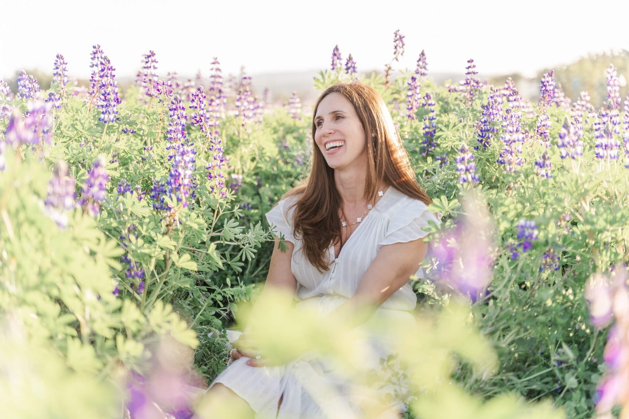 Brunette woman in white dress sitting in a green field of purple lupin flowers and laughing- Kindbody Newport Beach