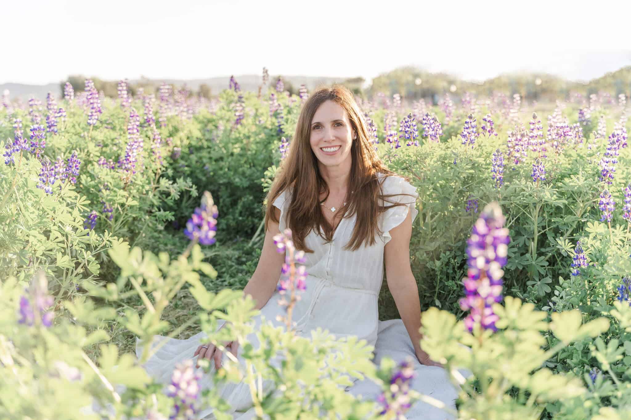 Smiling brunette woman in white dress sitting in a green field of purple lupin flowers