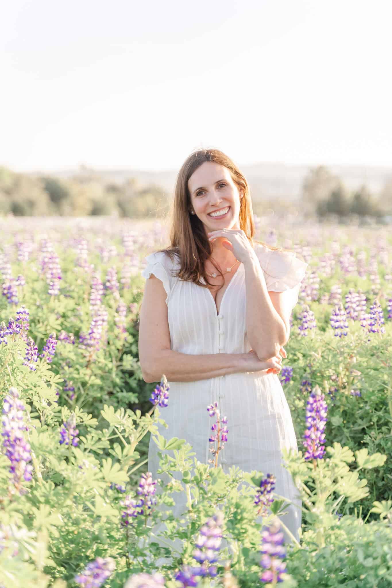 Brunette woman in white dress standing in a green field of purple lupin flowers- Kindbody Newport Beach