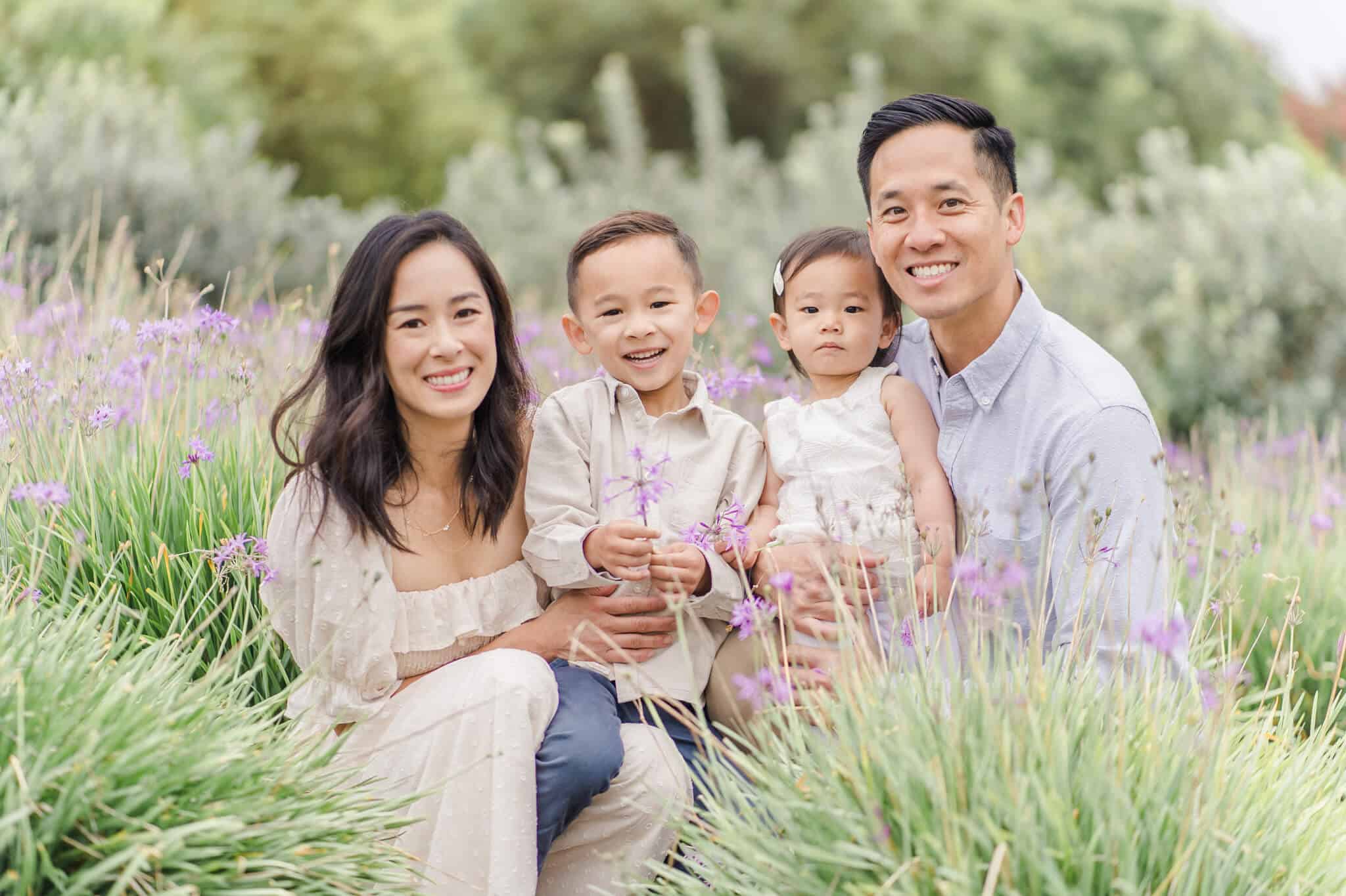Asian mom and dad holding toddler son and daugher in their arms while squatting in between some bushes/grass