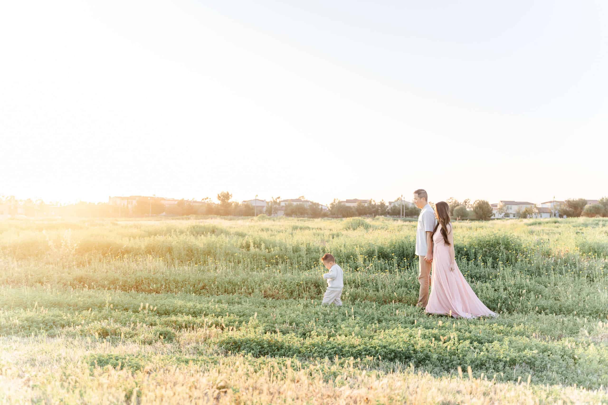 Mom in pink dress and dad walking behind toddler child in a field