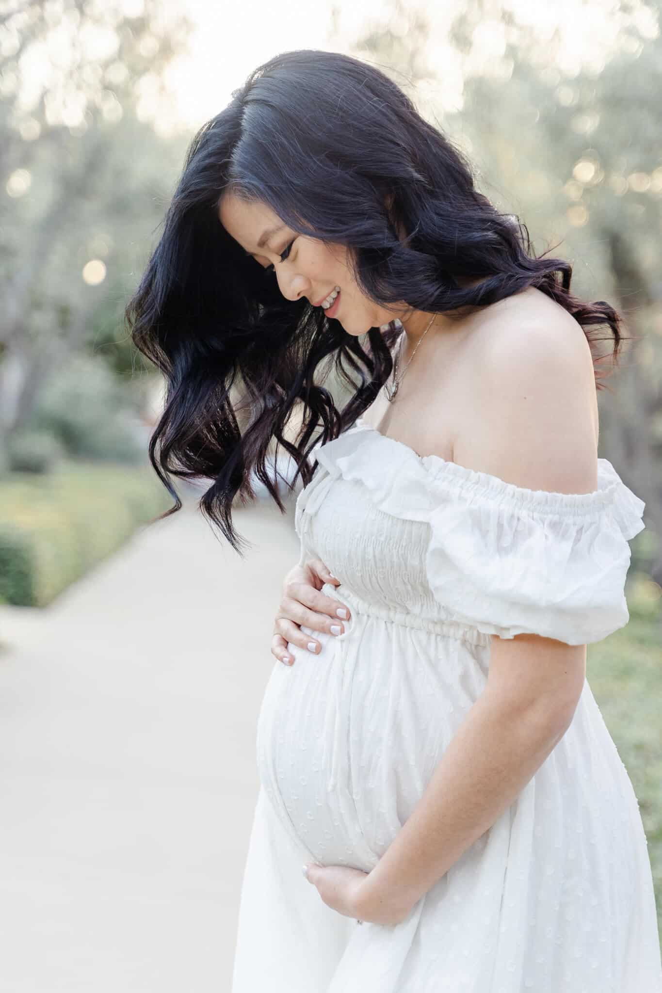 Pregnant mom looking down at her belly in white maternity dress. Junie Grey