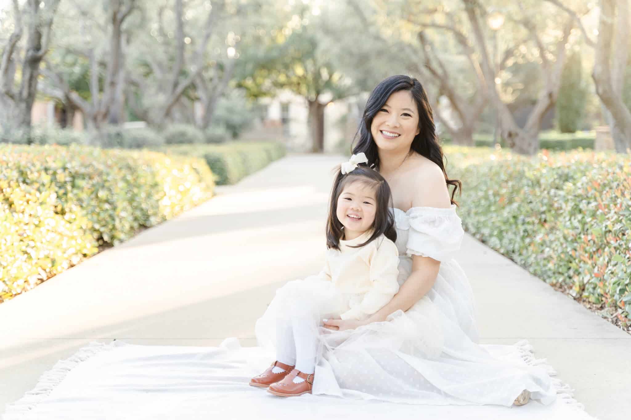 Pregnant mom in white dress and girl in cream dress sitting on her lap on white blanket. Junie Grey