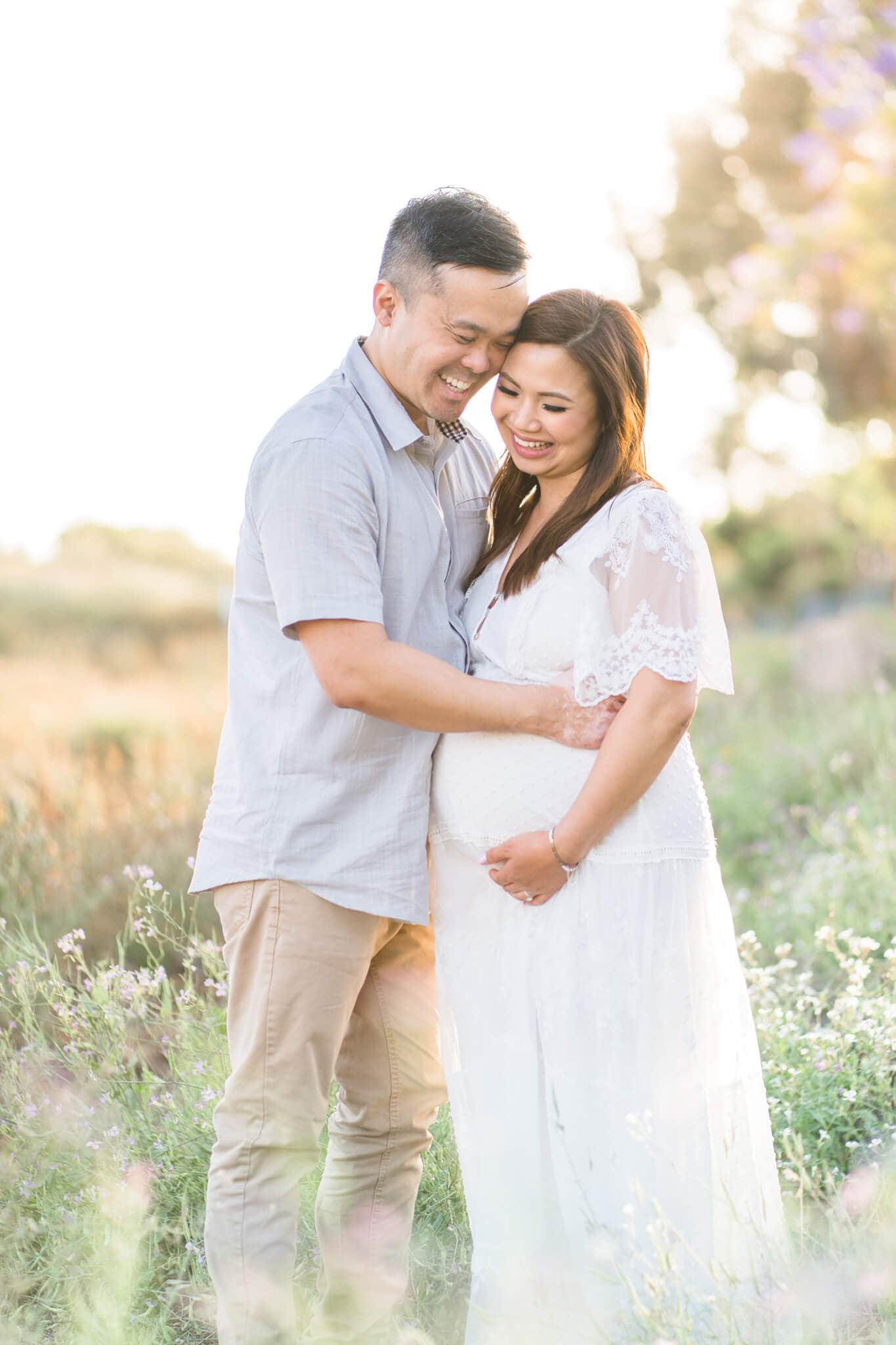 Pregnant mom and dad laughing and embracing each other on a grassy flower field. 