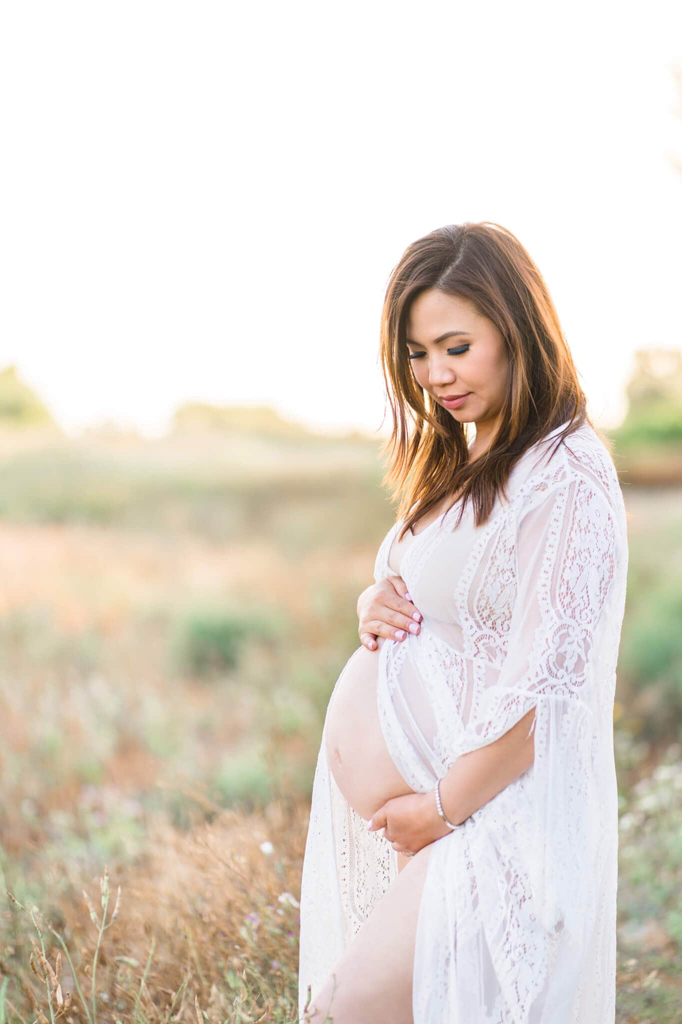 Pregnant mom in white dress showing her pregnant belly standing in a grassy field outdoors. Orange Coast Women's Medical Group