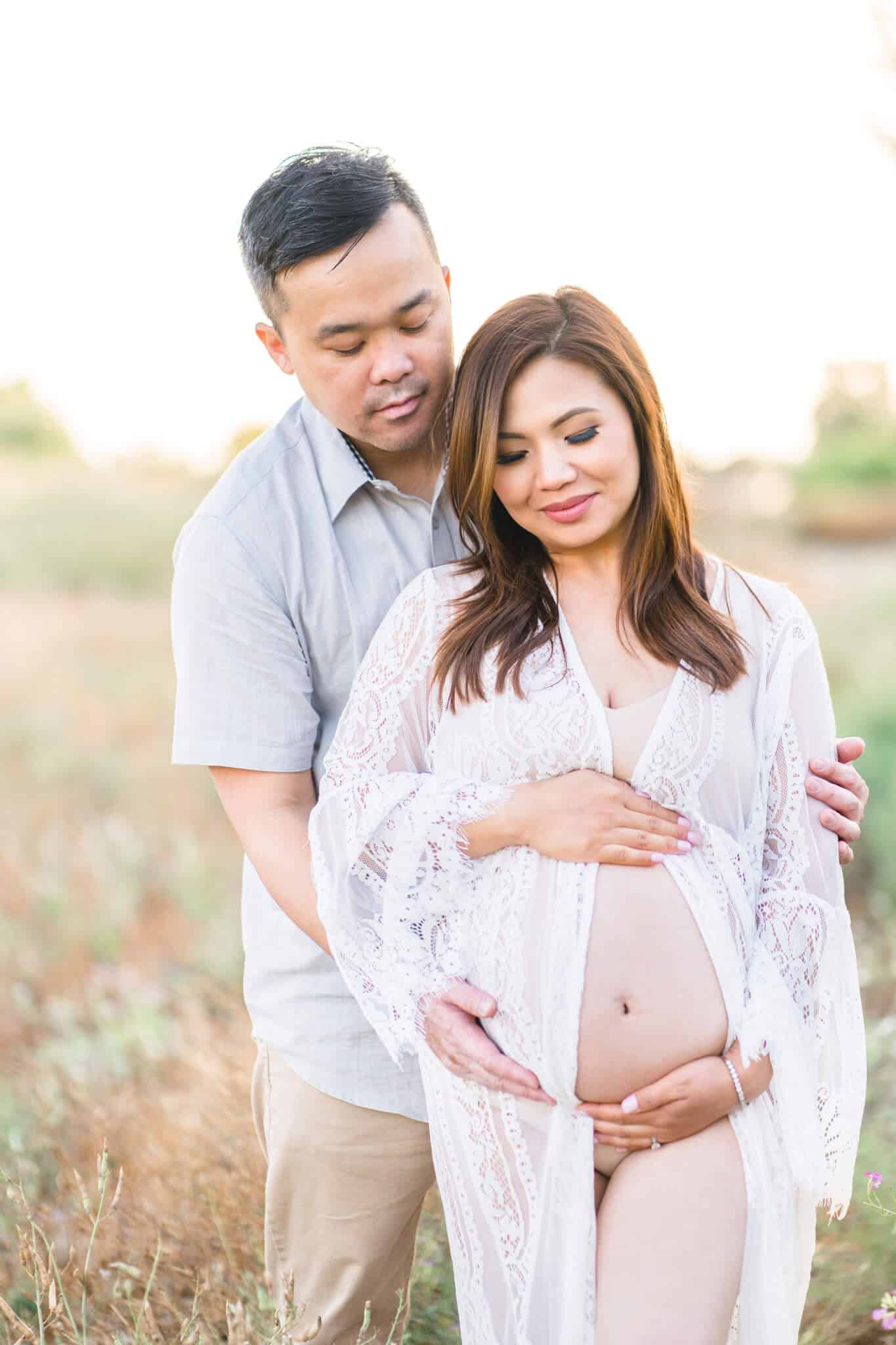 Pregnant mom holding her bare belly in a white dress outdoors in a field with dad standing and hugging her from behind. Orange Coast Women's Medical Group