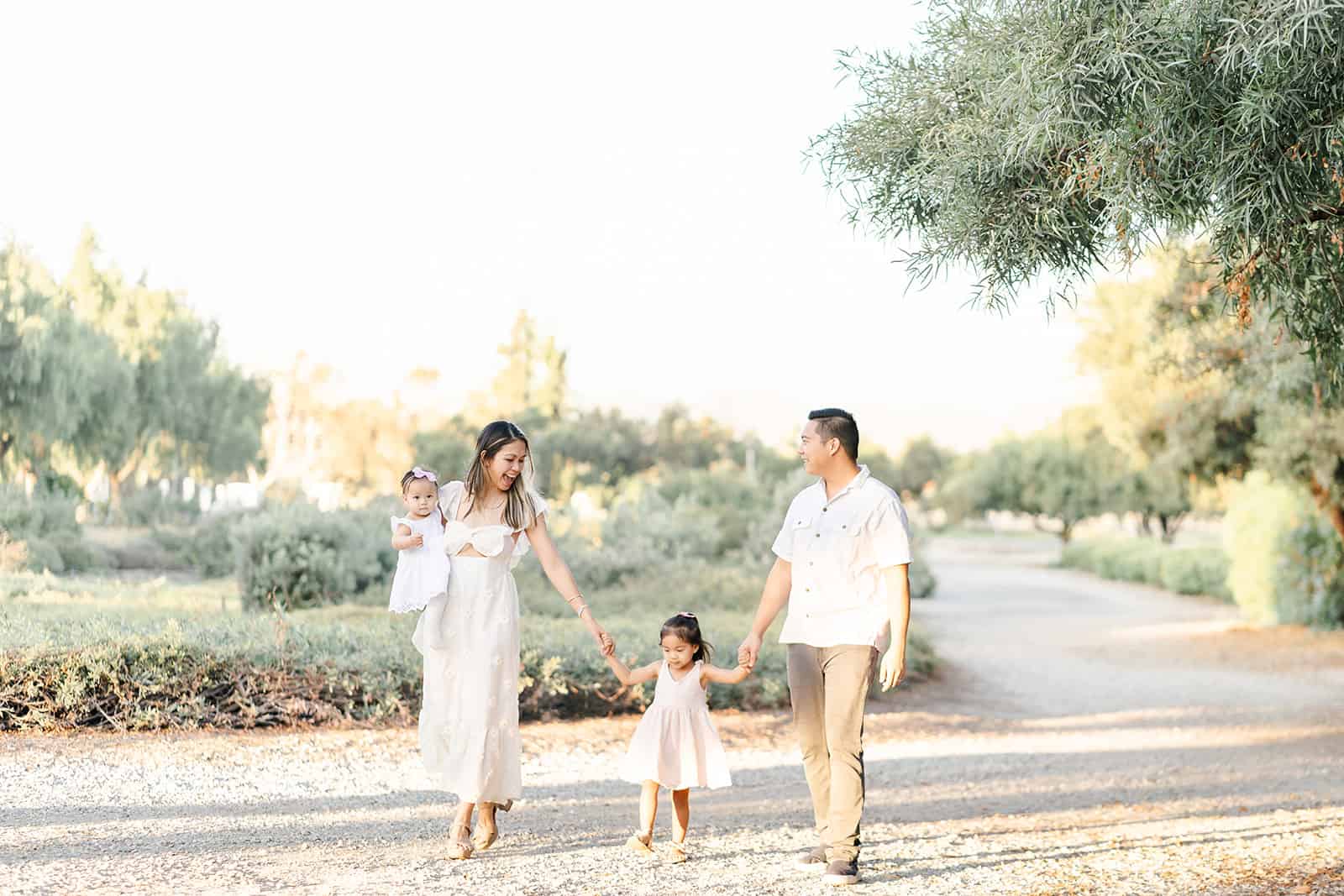 A mom and dad smile as they walk through a garden path holding hands with their toddler daughter as mom holds their infant daughter after attending Irvine International Academy