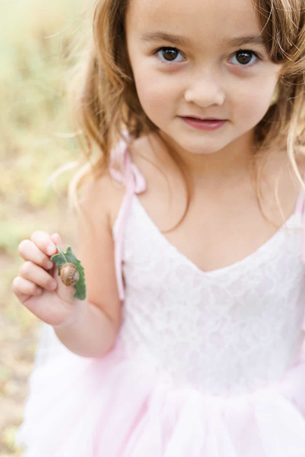 A young girl plays with a snail on a leaf while wearing a pink dress