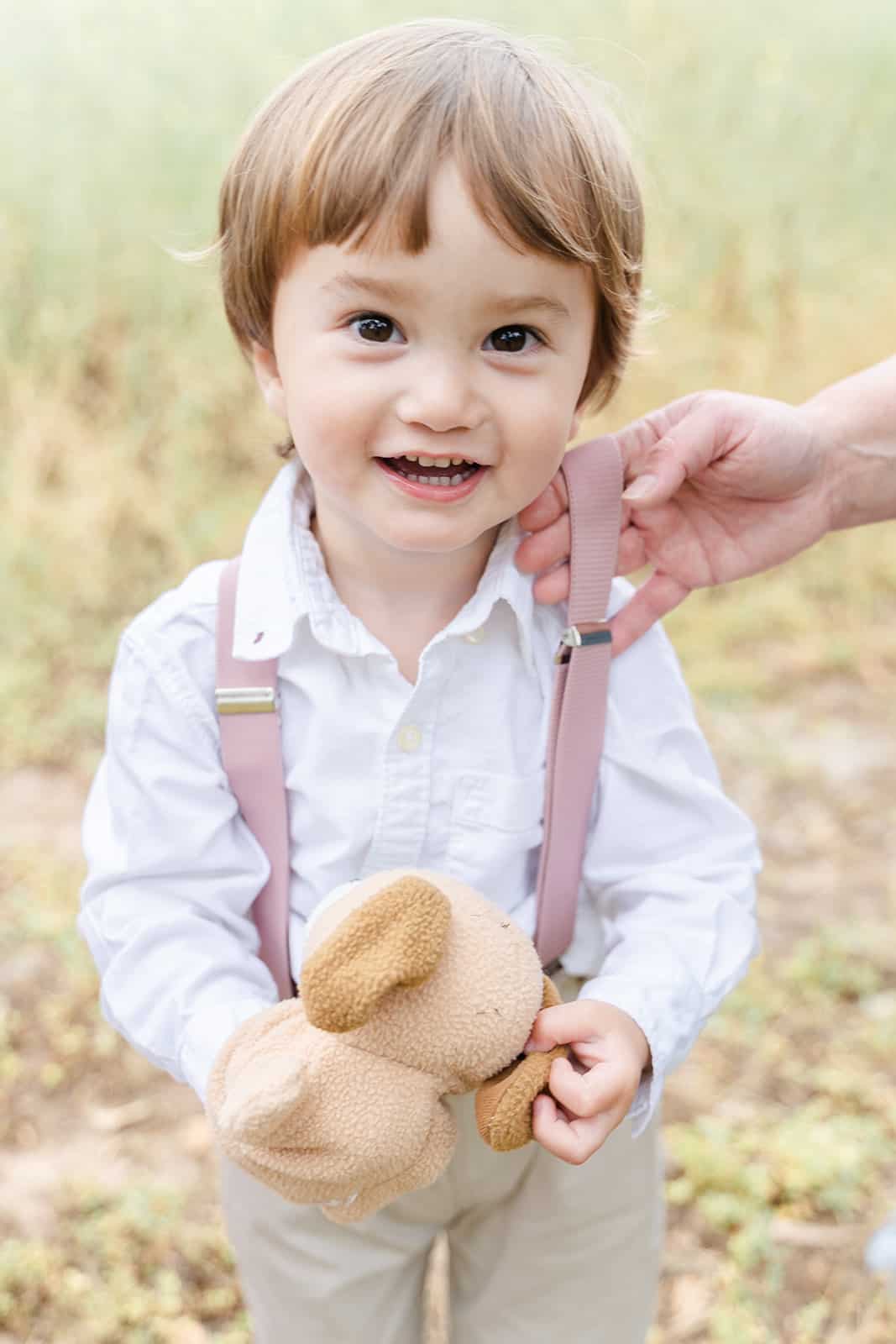 A young boy in pink suspenders plays with a stuffed dog in a field of tall grass