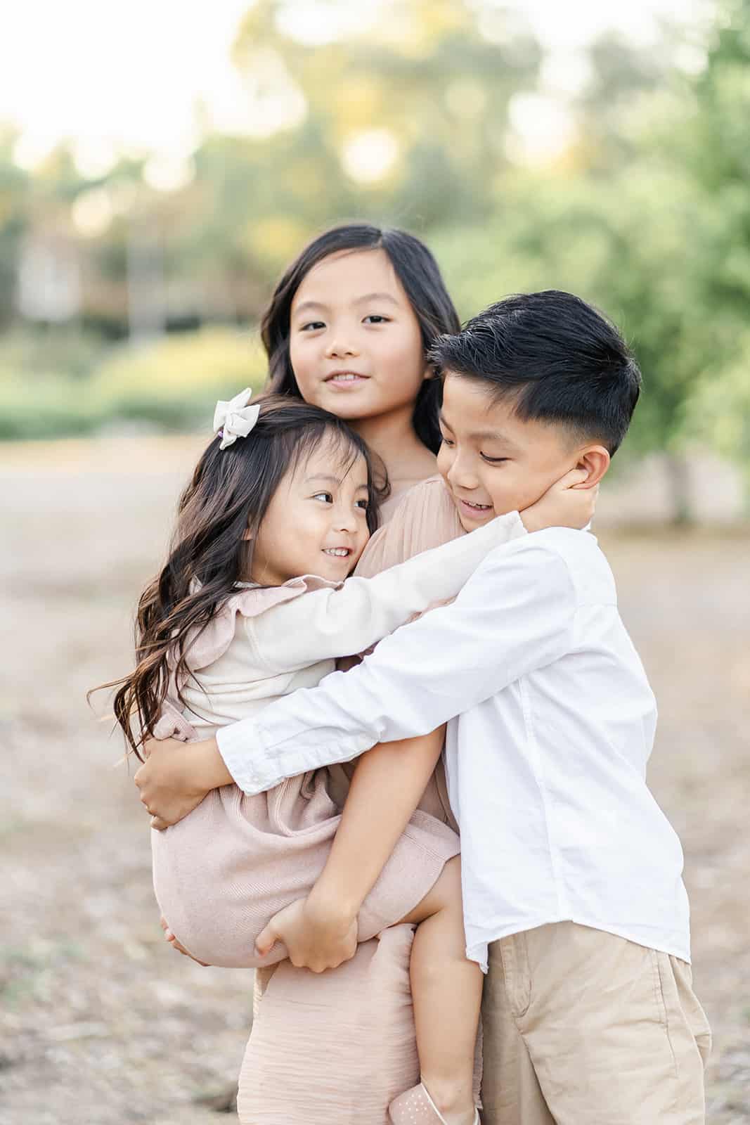 A young girl in a brown dress holds her toddler sister while being hugged by a brother in a park after attending Irvine Chinese School