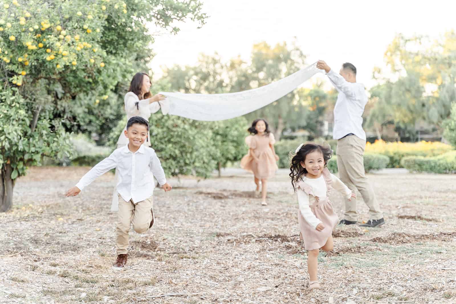 Mom and dad lift a blanket as their three Irvine Chinese School student children run underneath it in a park by an orange tree