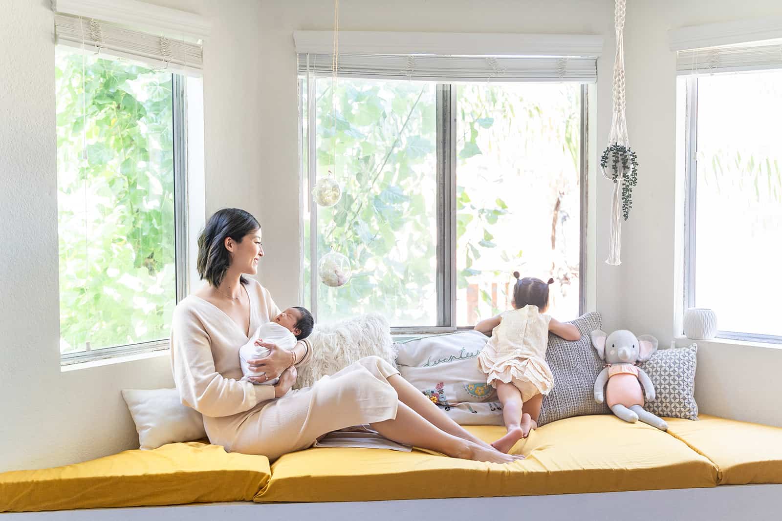 A mother sits along a bay window holding her sleeping newborn baby while her toddler daughter peeks out the window