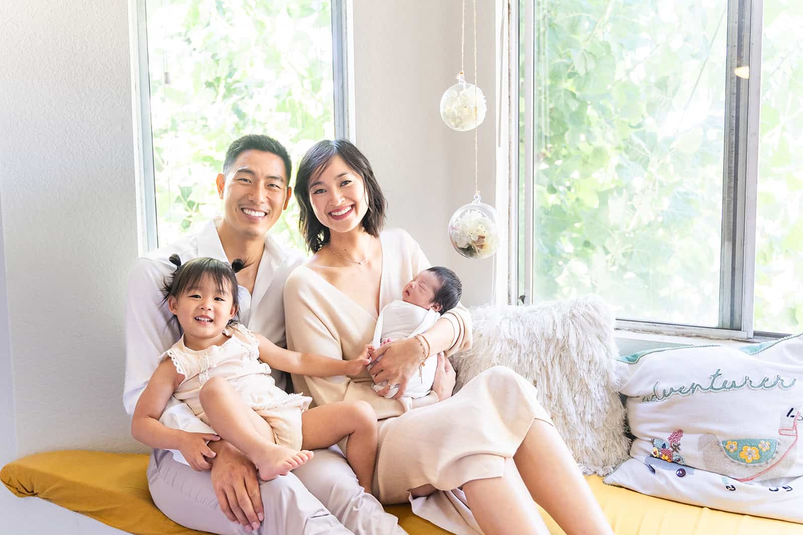 A mom and dad sit in a bay window with their newborn baby and toddler daughter in their laps