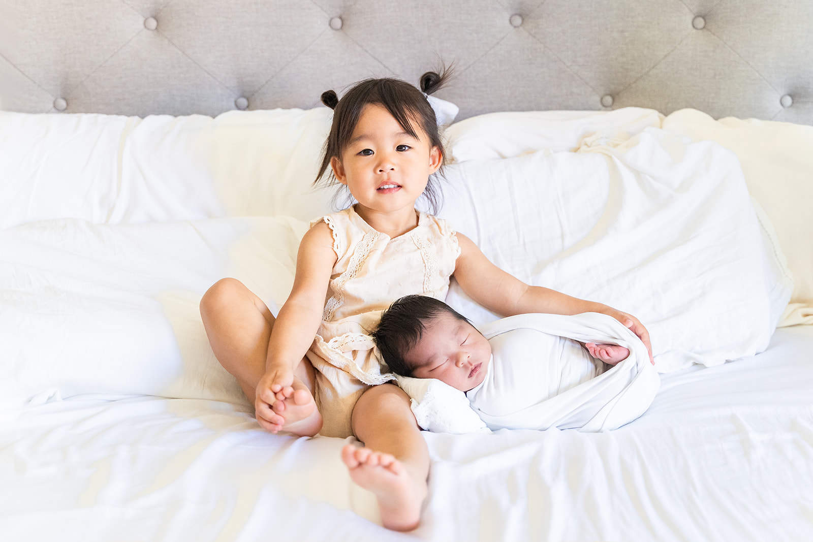 A young toddler girls in a white dress sits on a bed with an arm around her sleeping newborn sibling on her lap