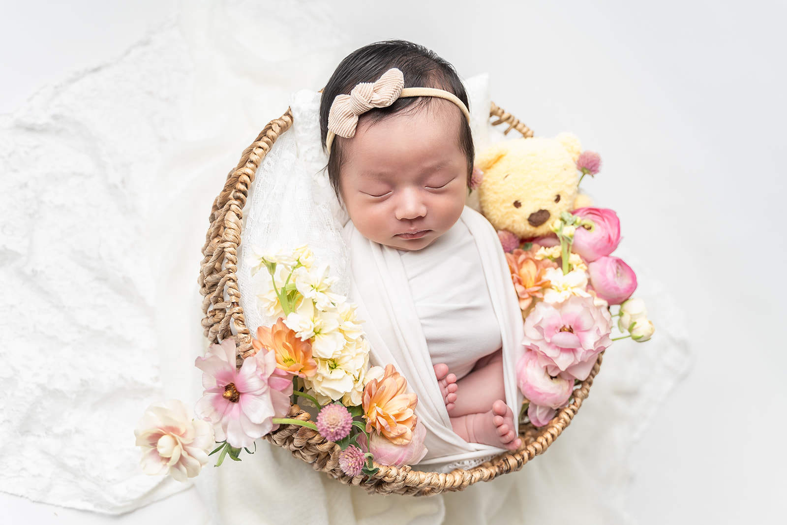A newborn baby girl in a small bow sleeps in a wicker basket with a bear and colorful flowers