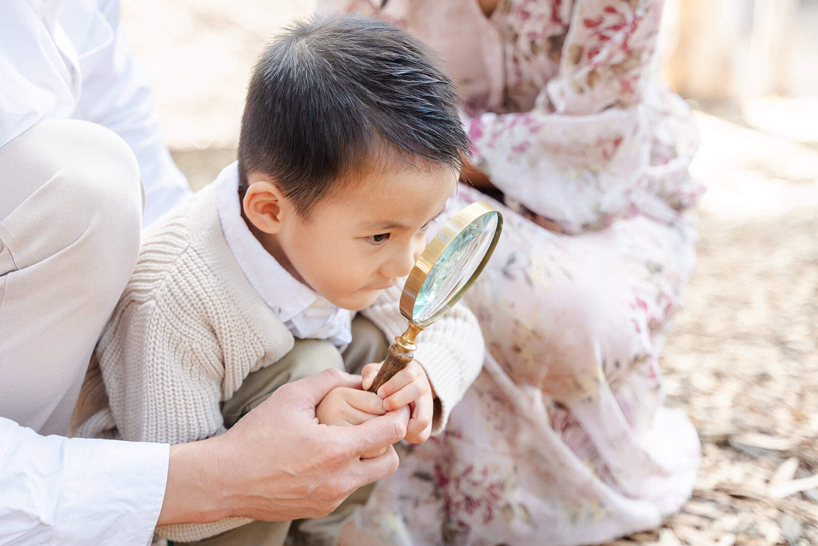 A young boy looks through a magnifying glass with help from mom and dad in a park