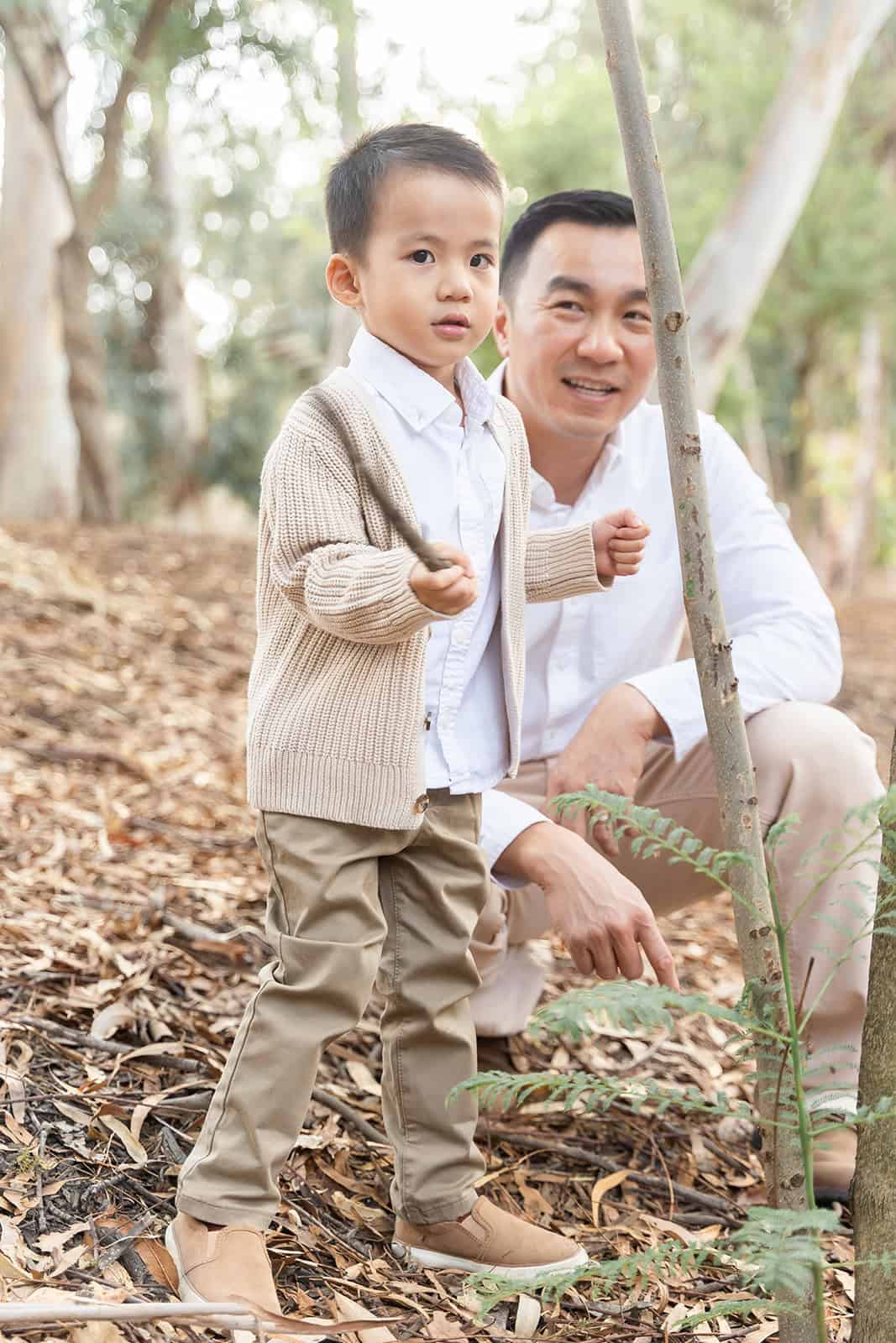A toddler boy in a tan sweater plays with a stick with dad in a park before visiting Irvine Regional Park