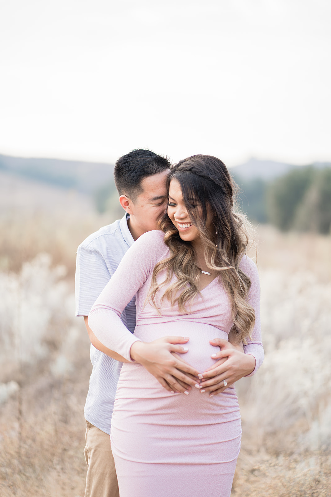 A mom to be holds her bump with her husband while being hugged from behind in a field of tall grass thanks to Life IVF Irvine