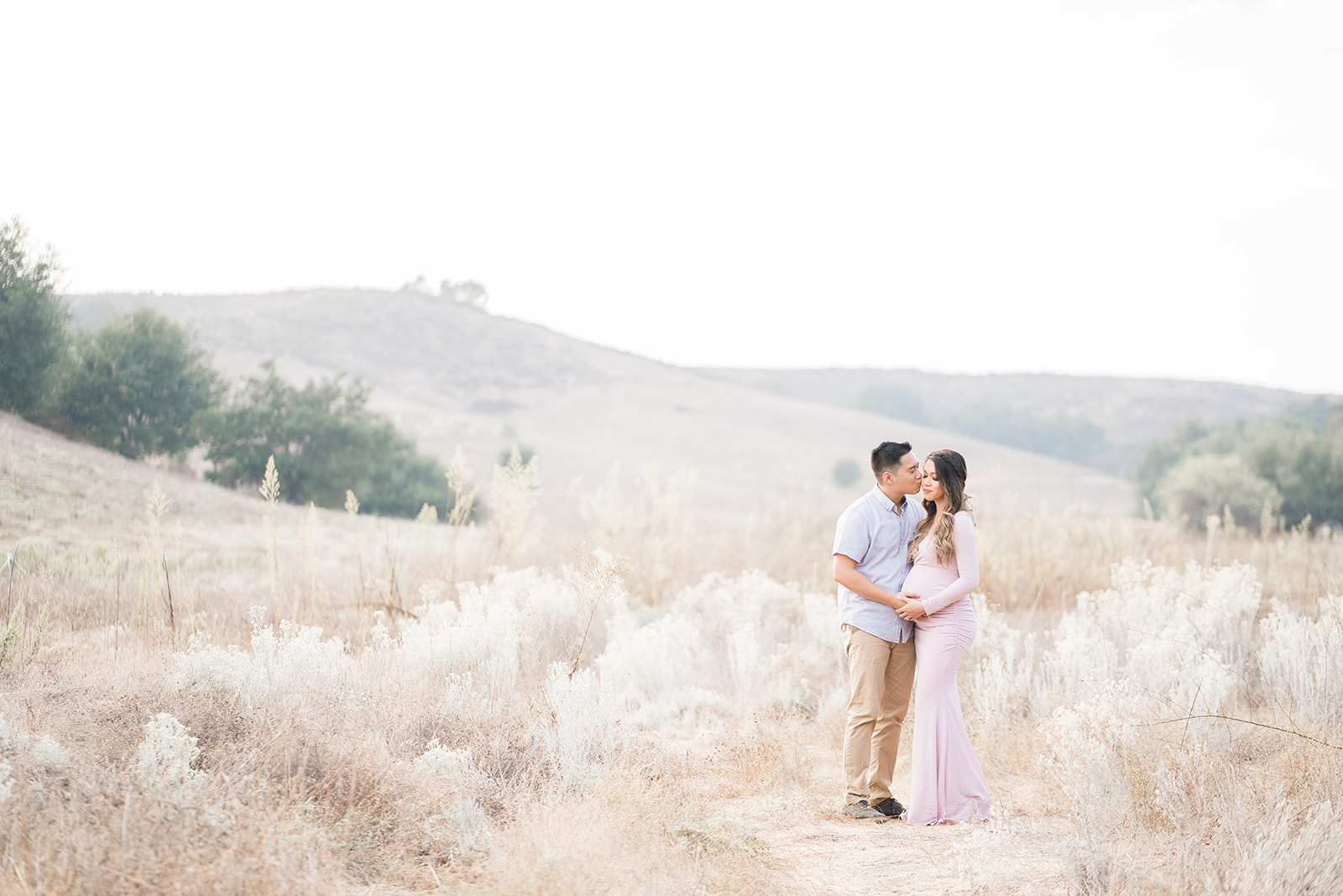 Expecting parents stand in a field of tall golden grass while sharing an intimate moment with hands on the bump