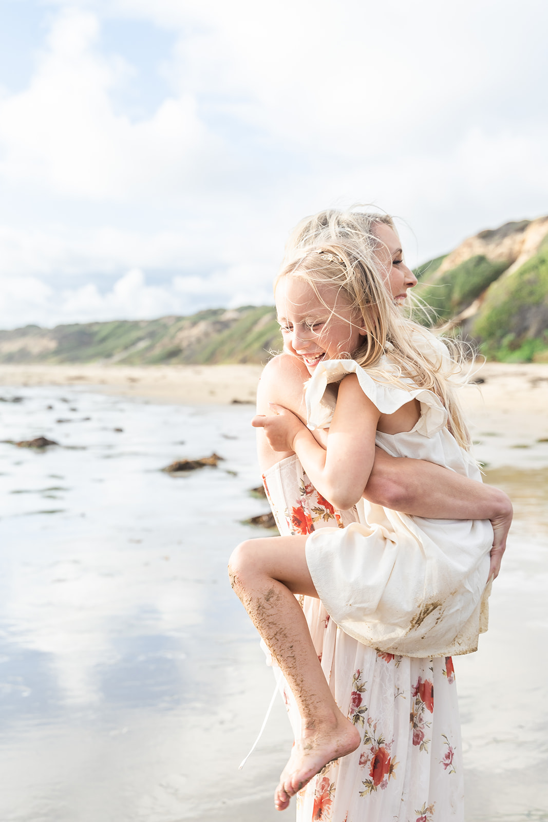 A mother in a floral dress carries her daughter on her hip while standing on a beach