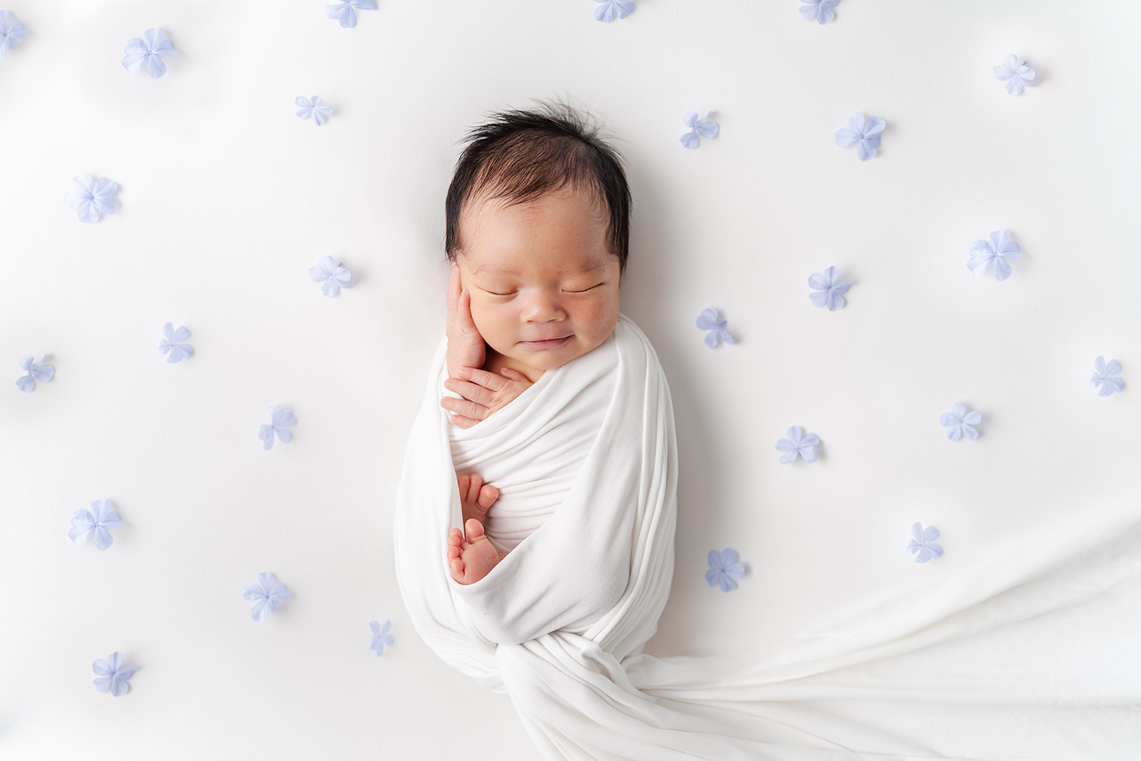 A newborn baby sleeps in a white swaddle surrounded by blue flowers in a studio