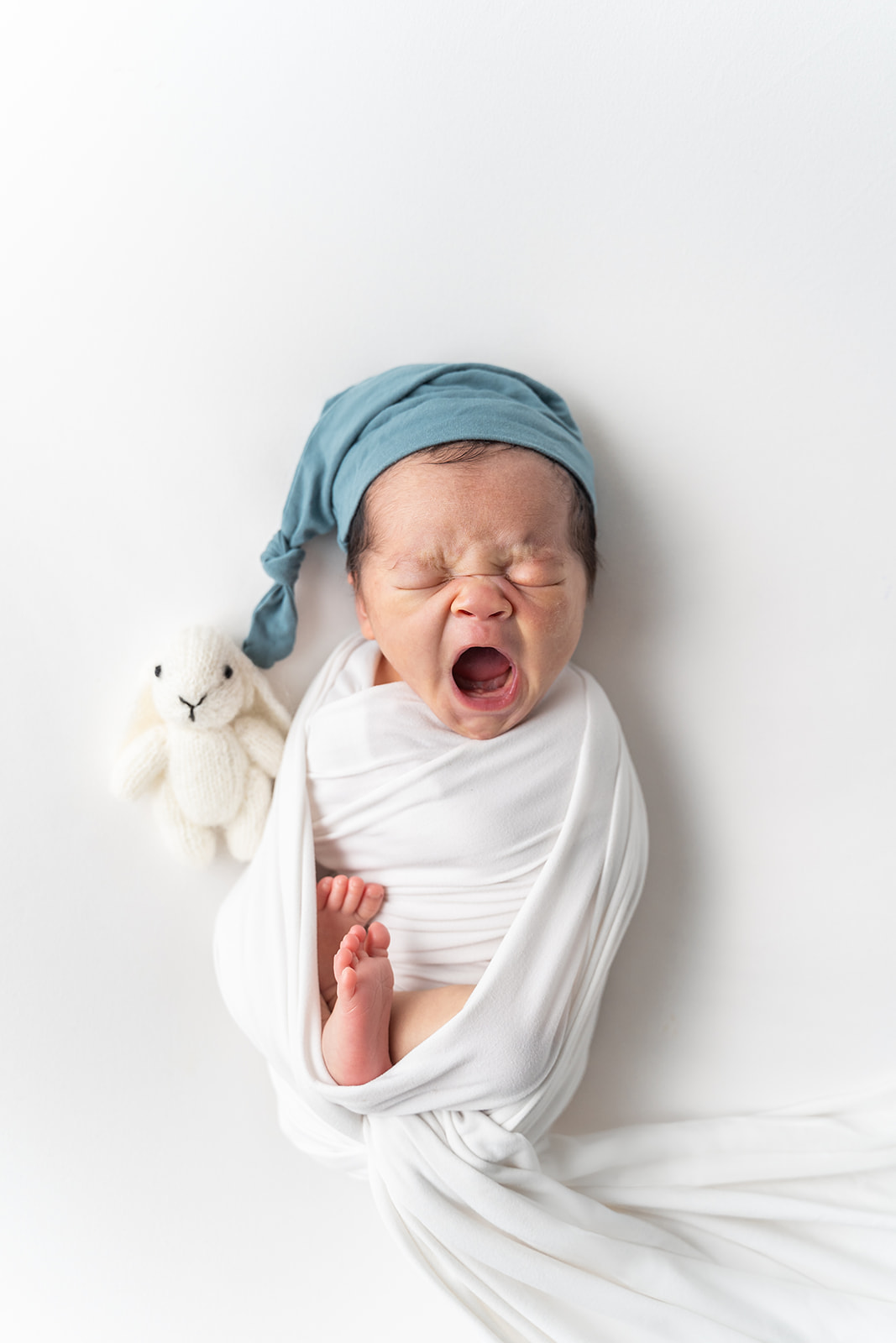 A newborn baby in a blue sleep cap yawns while laying on a bed in a studio