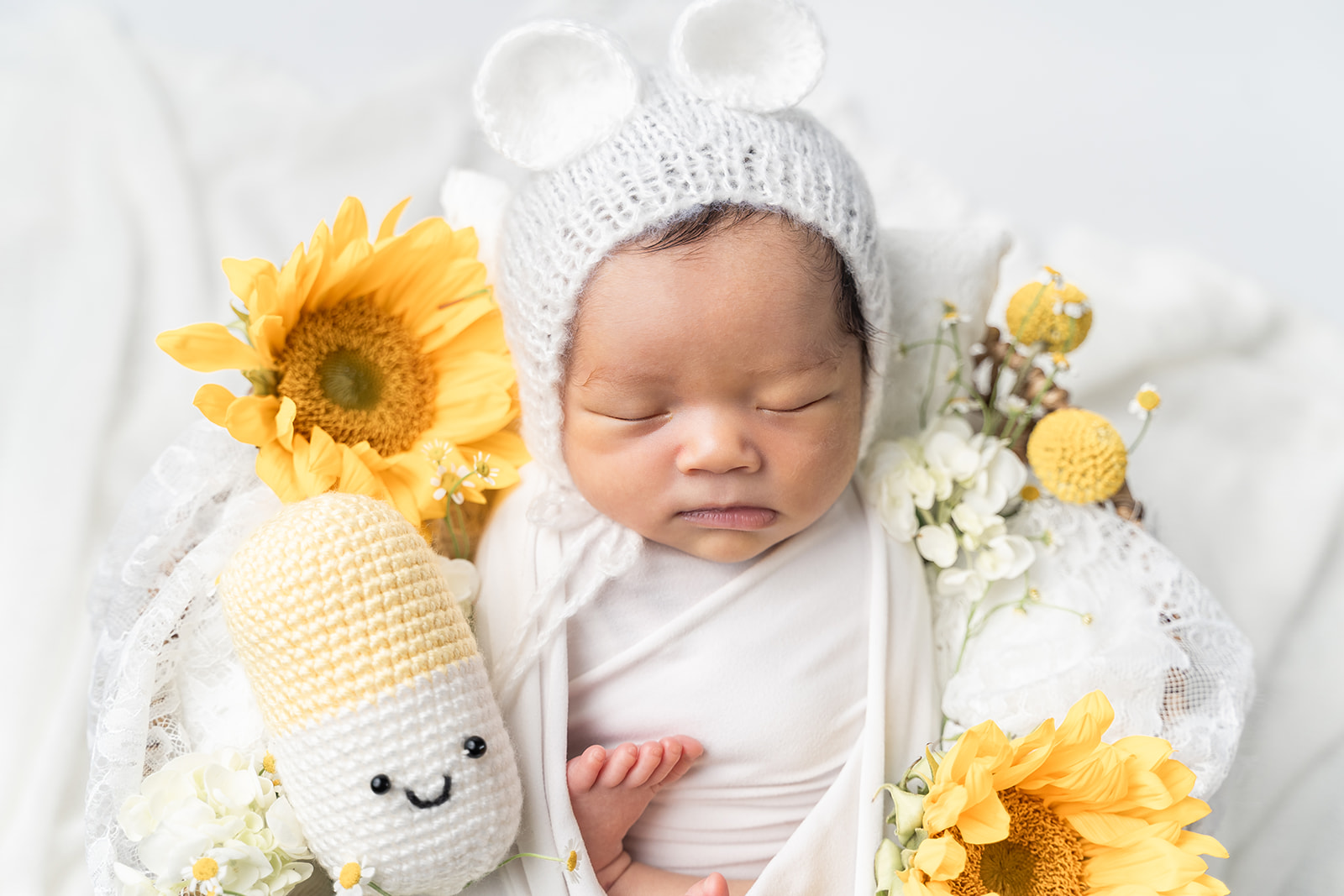 A newborn baby in a knit bonnet with ears sleeps in a basket with sunflowers and other wildflowers in a studio with help from South Coast Midwifery