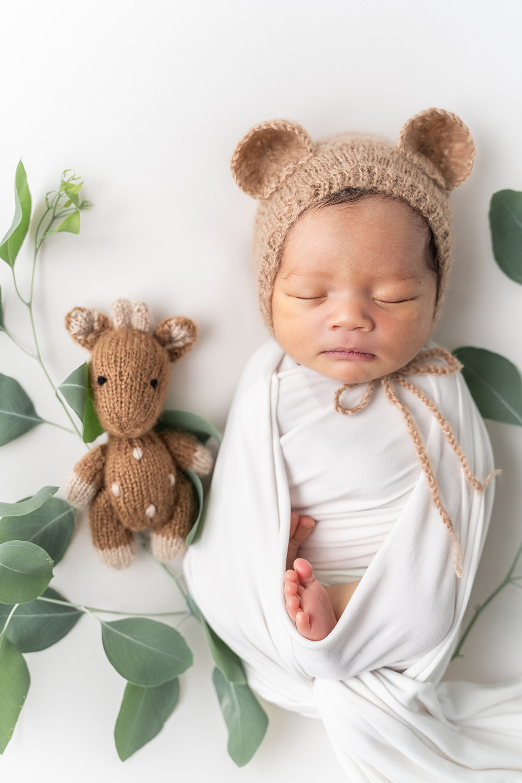 A newborn baby sleeps with a knit giraffe in a brown bonnet with ears