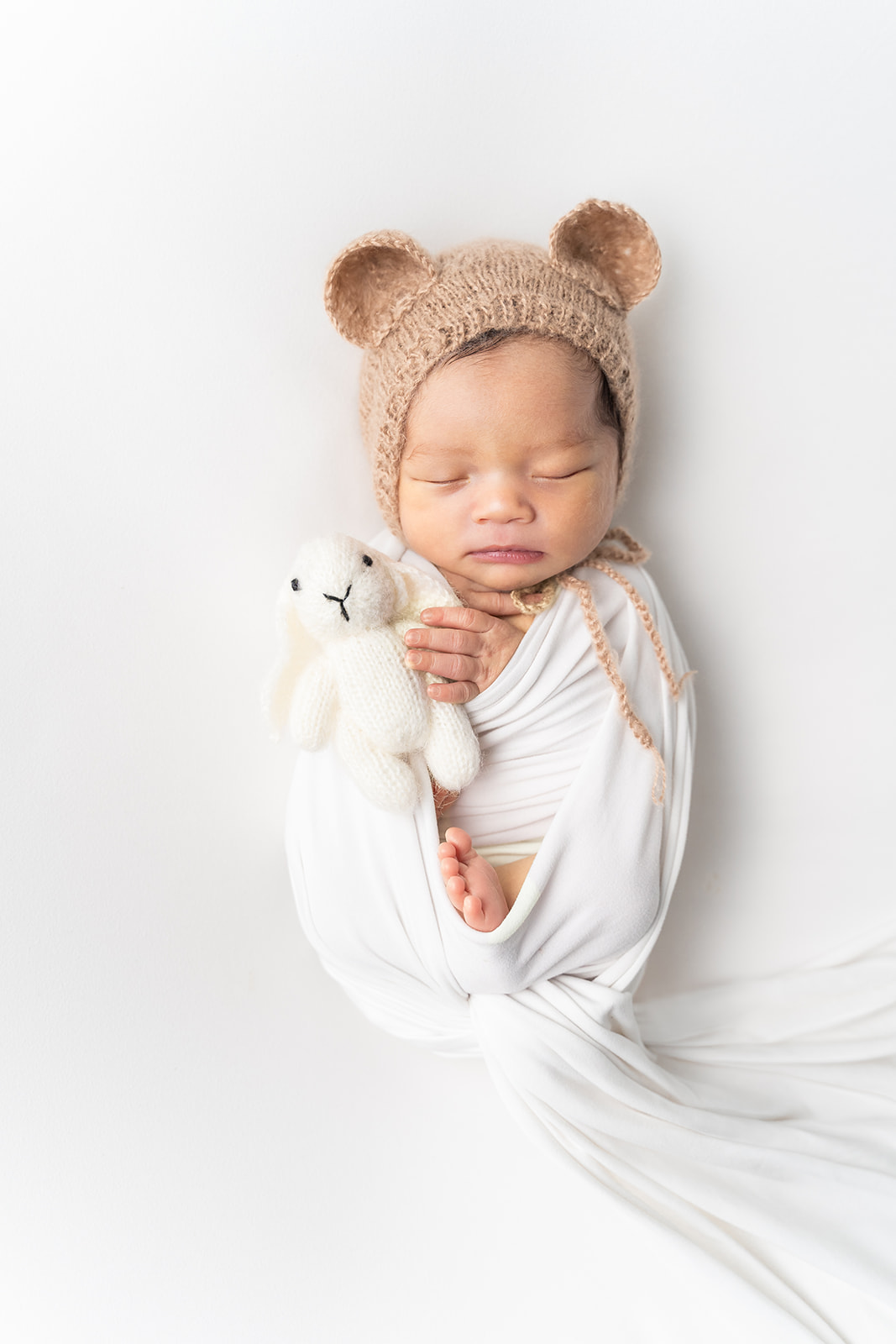 A newborn baby sleeps holding a white knit bunny in a bonnet with ears in a studio after visiting After The Stork