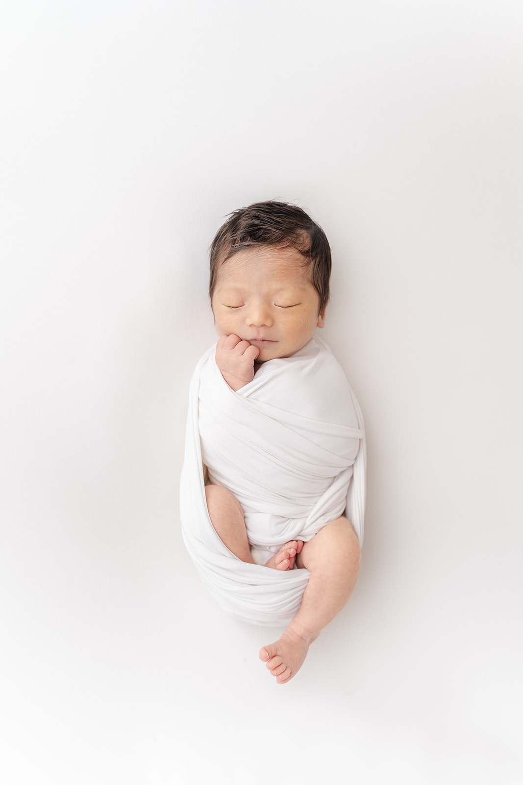A newborn baby sleeps with one leg sticking out of a white swaddle in a studio