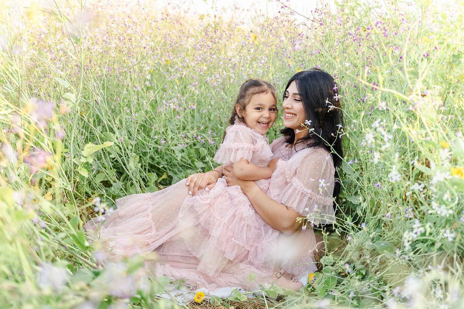 A young girl in a pink dress sits on mom's lap laughing surrounded by wildflowers