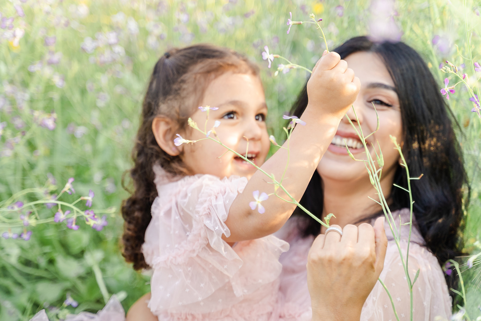 A mother and daughter in matching pink dress play with flowers while sitting in a field of wildflowers