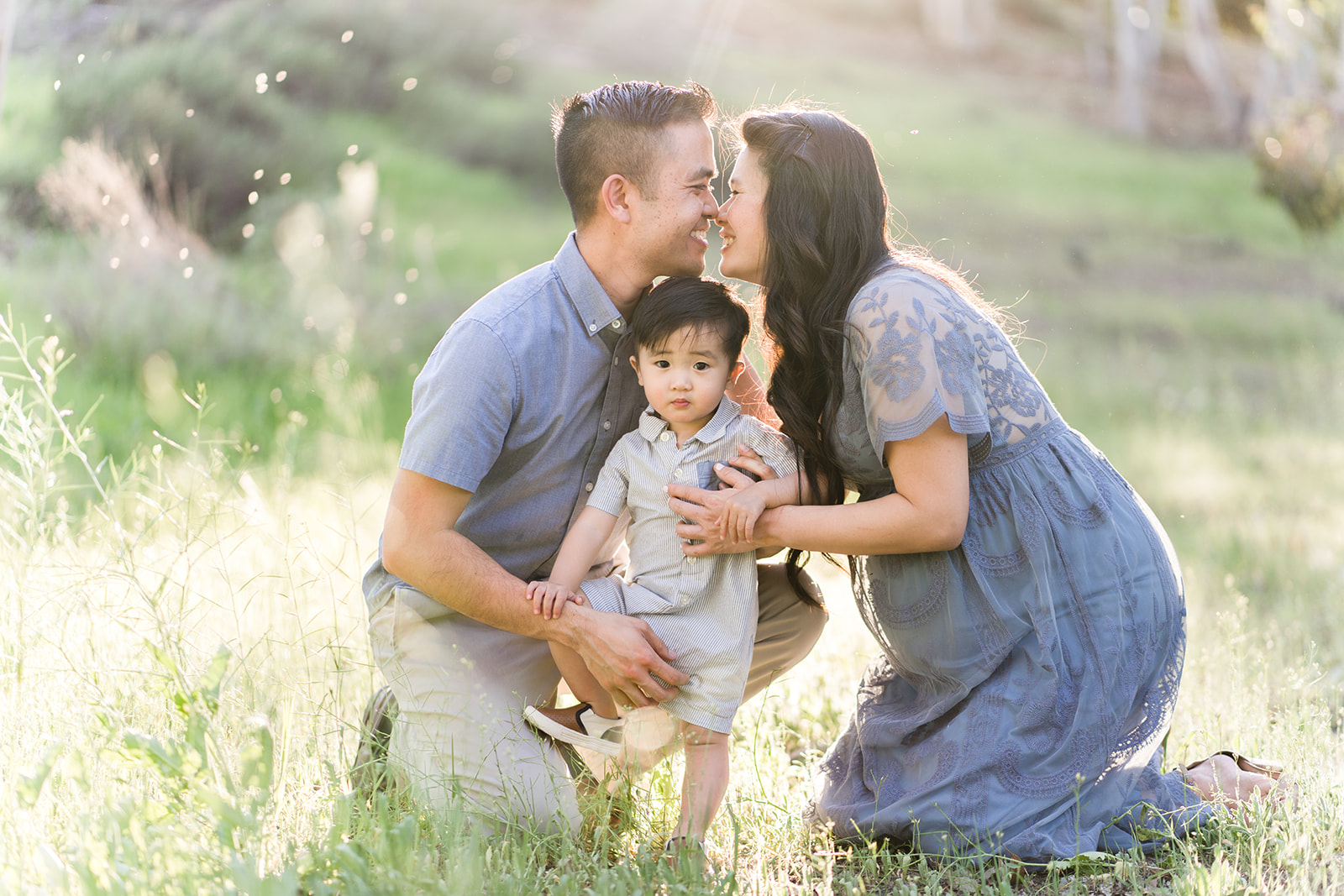 A mom and dad lean in for a kiss while kneeling over their toddler son in a field at sunset