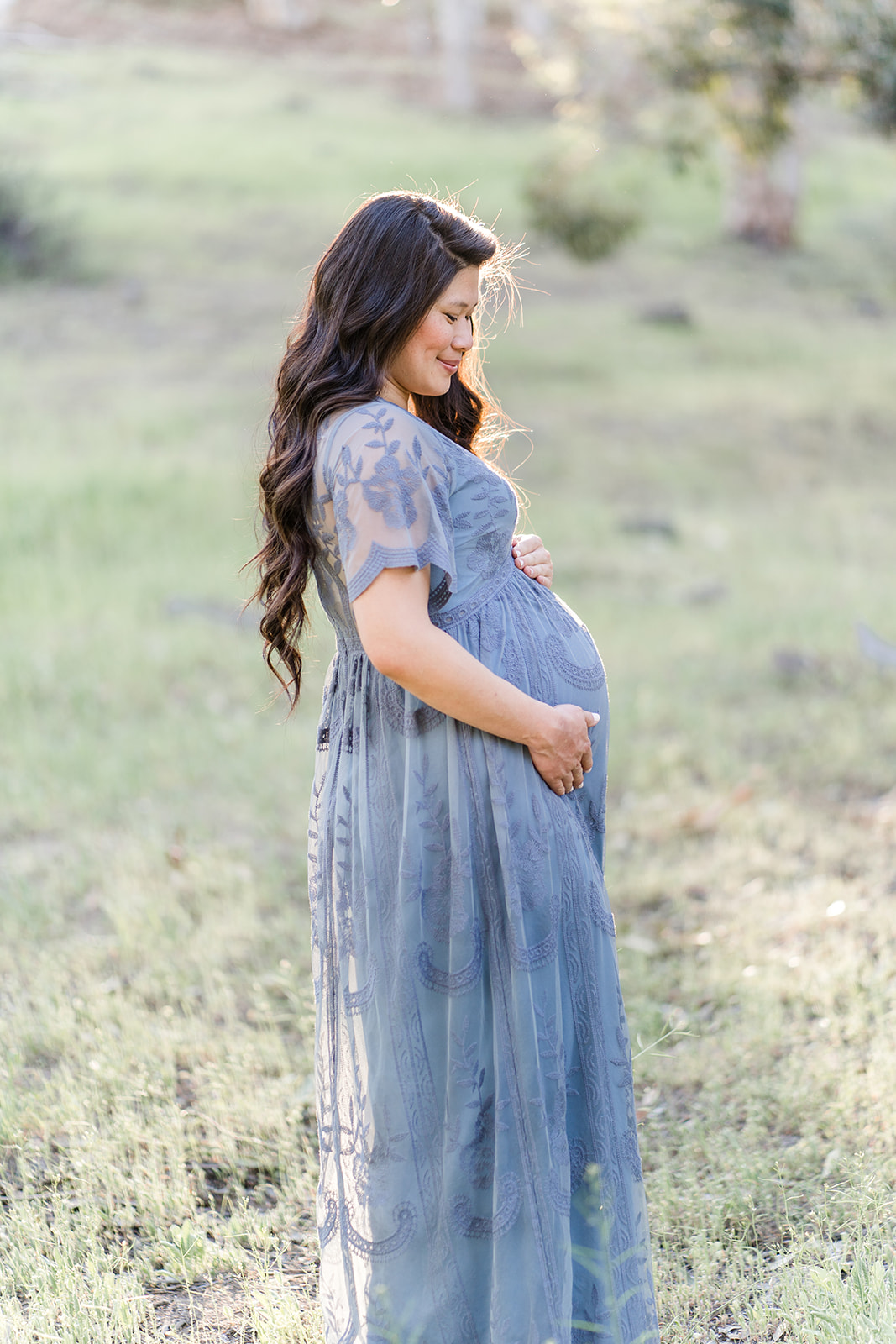 A mom to be in a blue lace dress stands in a field at sunset after visiting a Hoag OBGYN