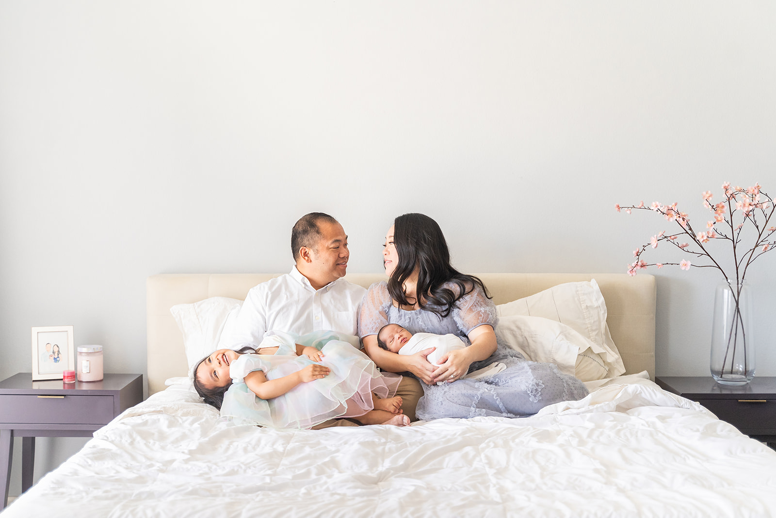 A mom and dad lean on each other while sitting on a bed with their newborn baby and toddler in their laps