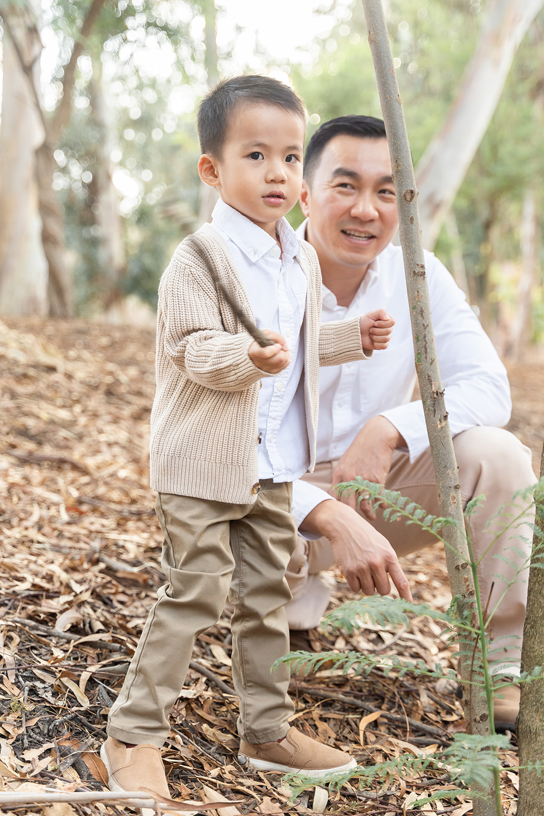 A toddler boy in a tan sweater plays with a stick with dad in a park before visiting Irvine Park Railroad