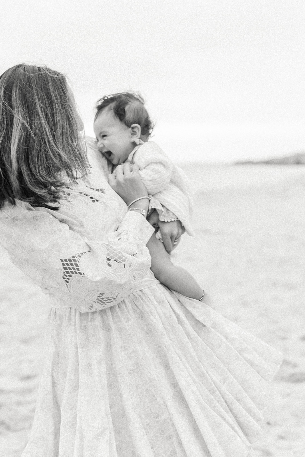 A mom in a lace dress twirls on a beach with her infant baby laughing in her arms before visiting Newport Montessori School