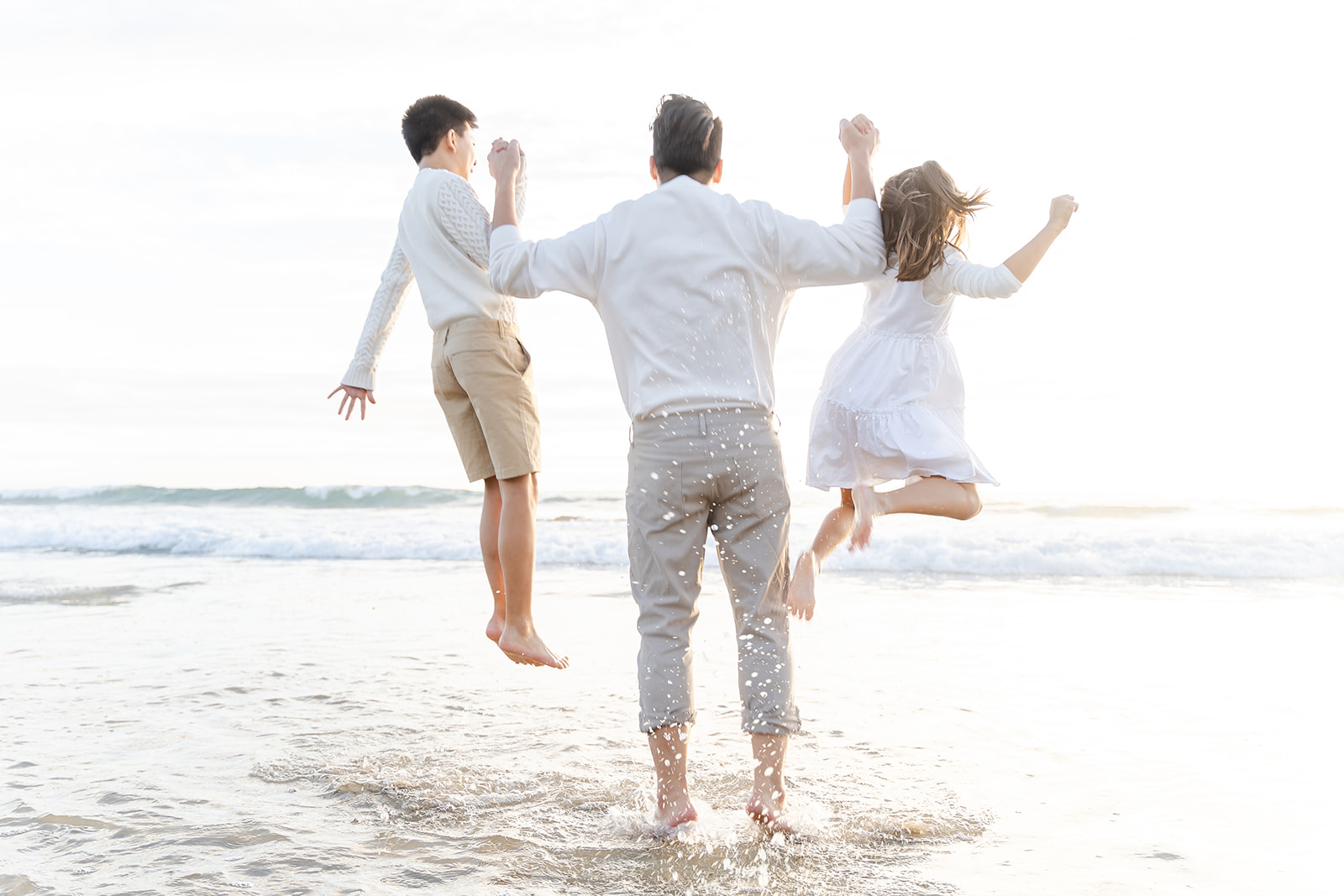 A father in a white shirt and khakis lifts his young son and daughter high up on the beach
