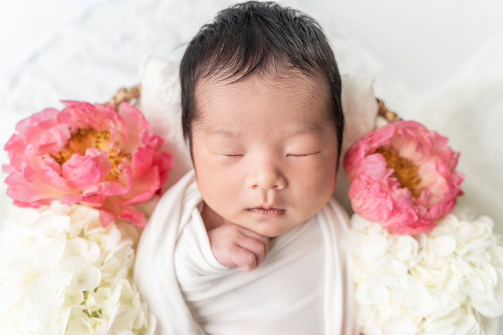 A newborn baby sleeps with a hand under her chin with pink carnations