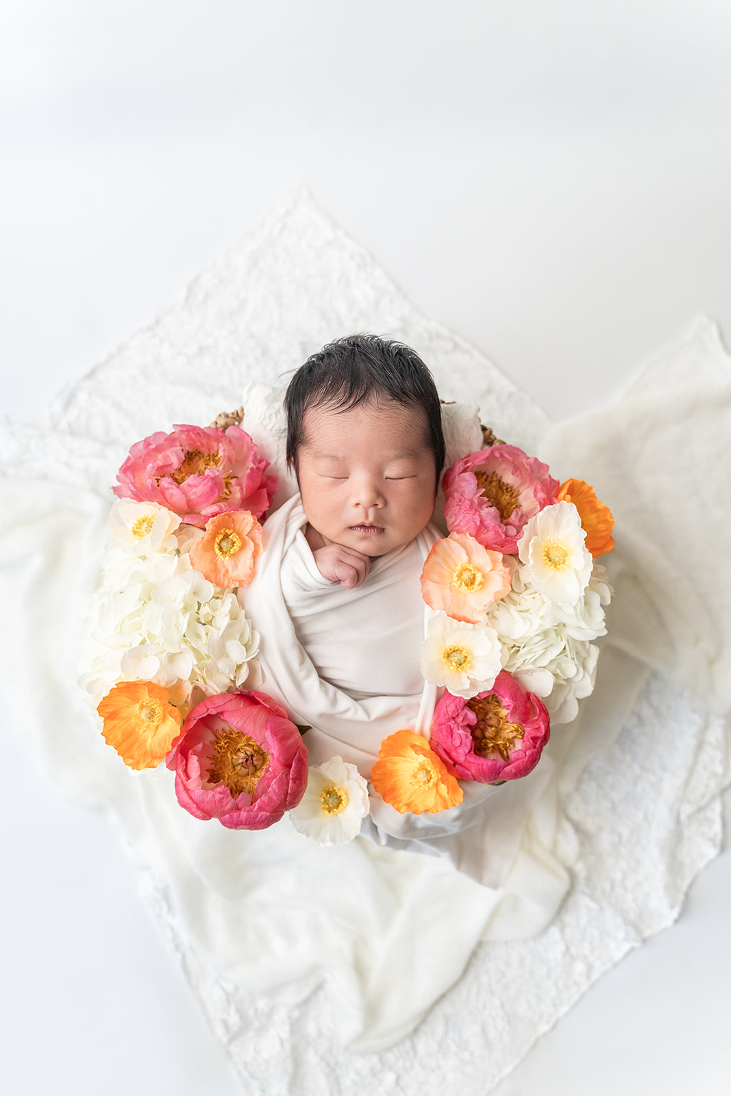 A newborn baby sleeps in a basket filled with colorful pink flowers in a studio