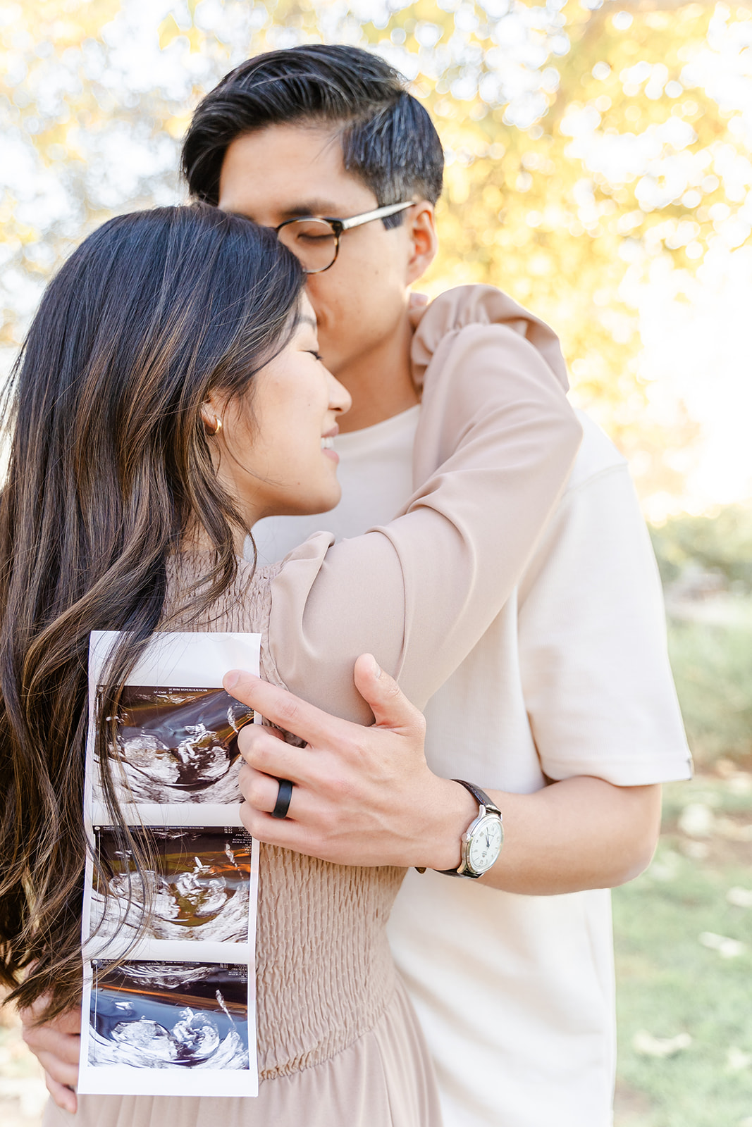 Expecting parents hug while holding the sonogram in a park at sunset thanks to an OBGYN Orange County