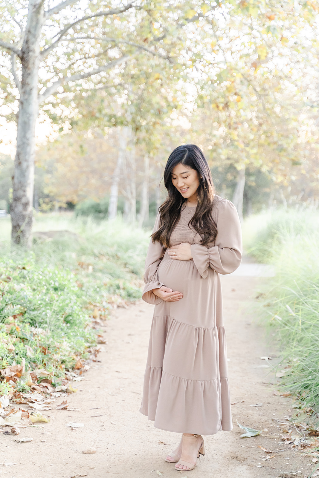 A mom to be in a brown maternity dress stands in a park path smiling down to her bump after visiting an OBGYN Orange County