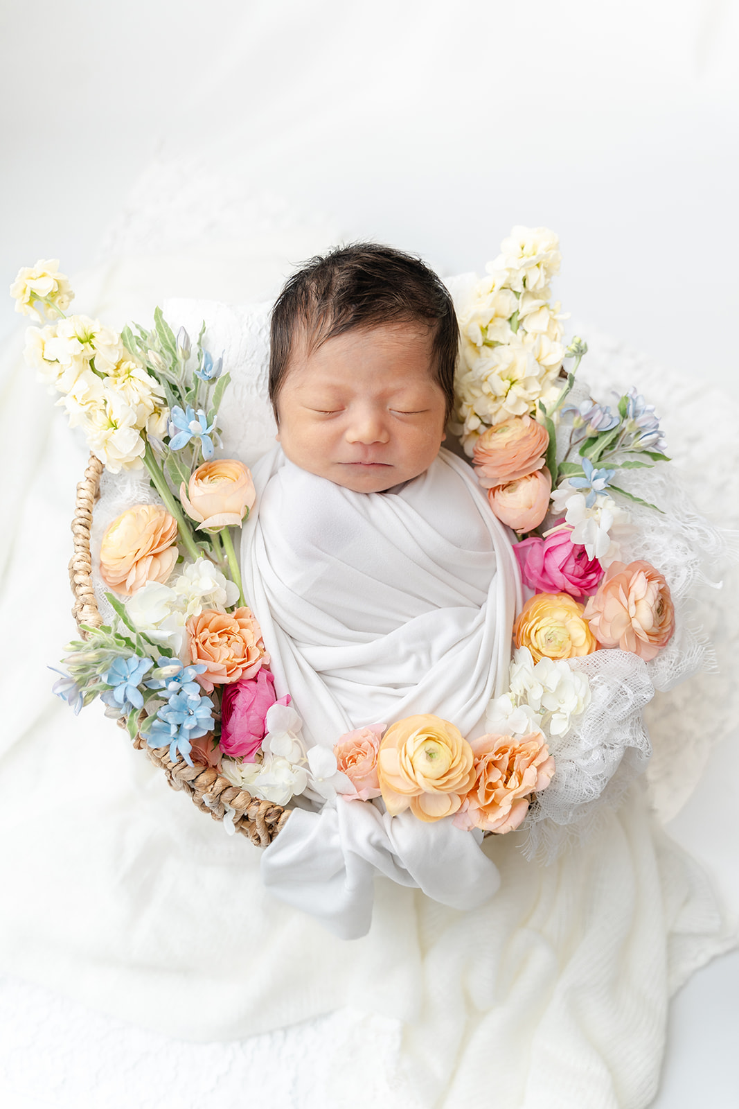 A newborn baby sleeps in a white swaddle in a woven basket surrounded by flowers