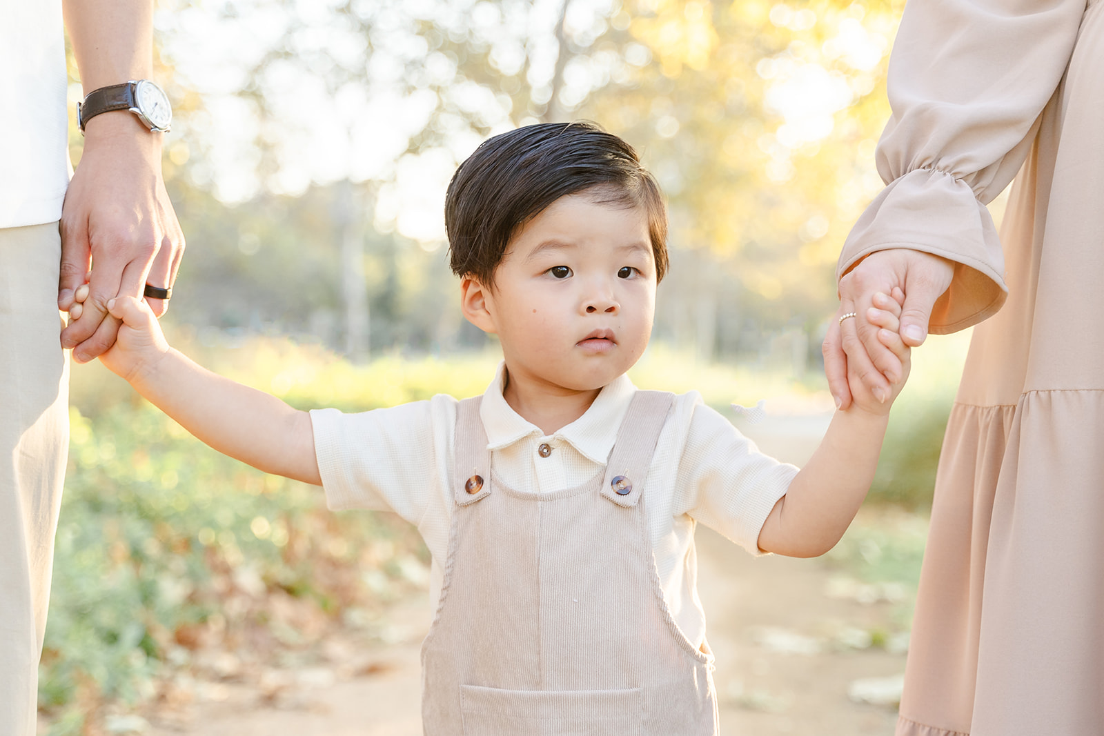 A young boy in brown overalls walks through a park holding hands with mom and dad after attending Preschool in Irvine